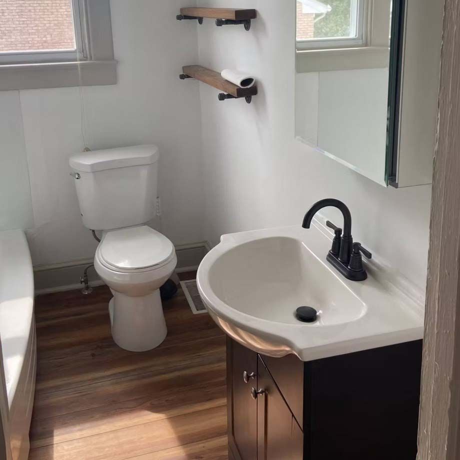 Small bathroom with toilet, pedestal sink, dark wood vanity, white walls, and window light.
