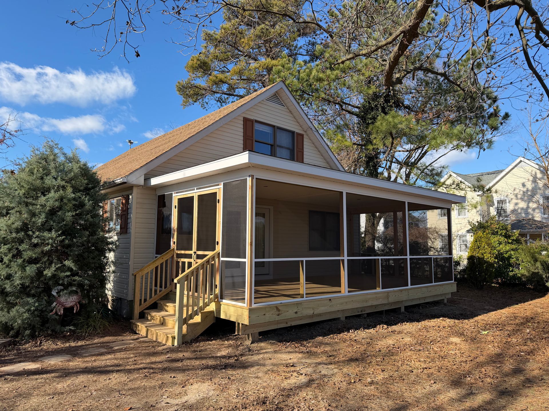 Small beige house with a screened porch, front steps, and a large tree in the yard under a blue sky.