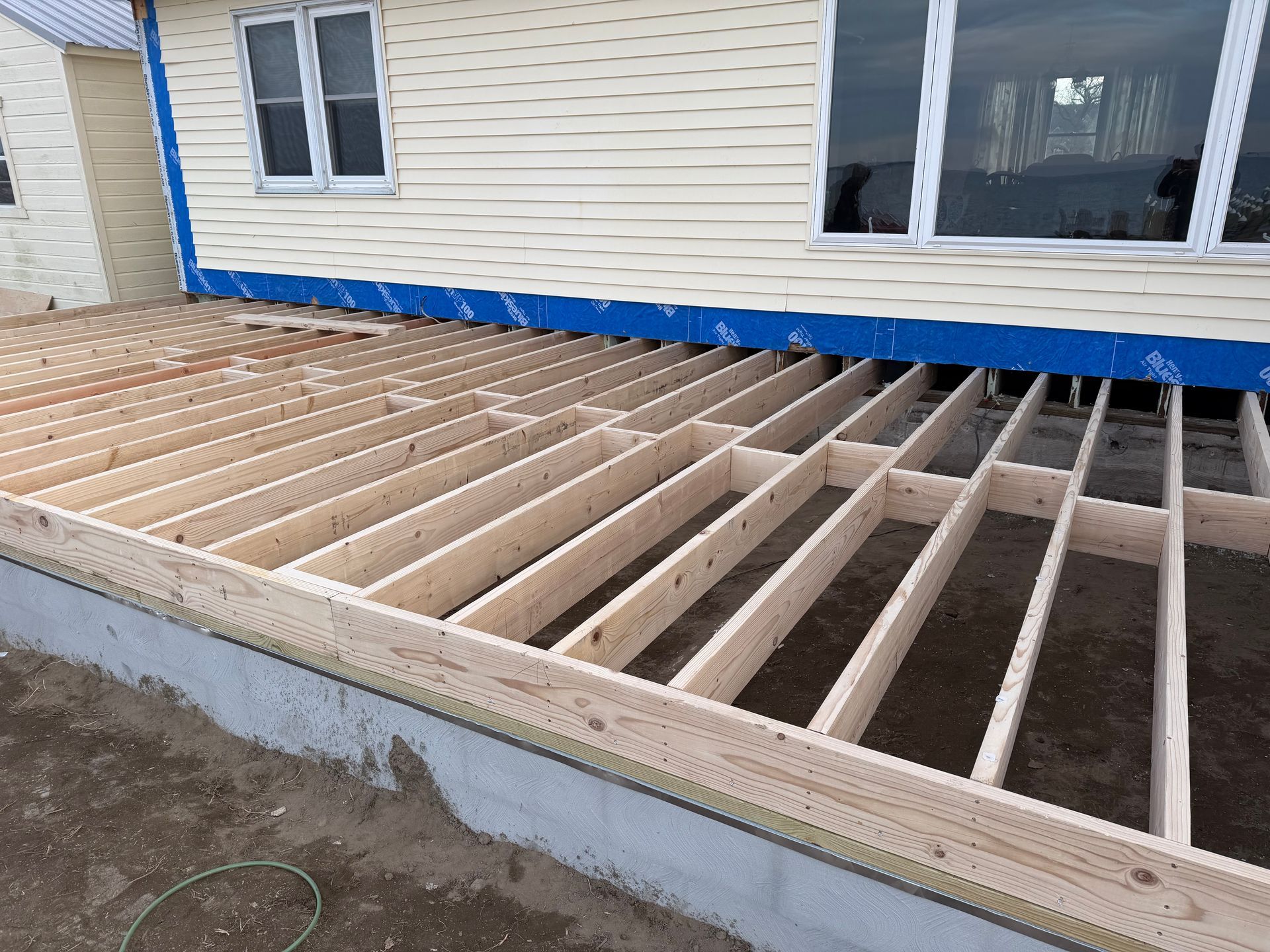 Partially built deck frame beside a beige house, with exposed wooden joists and blue trim.