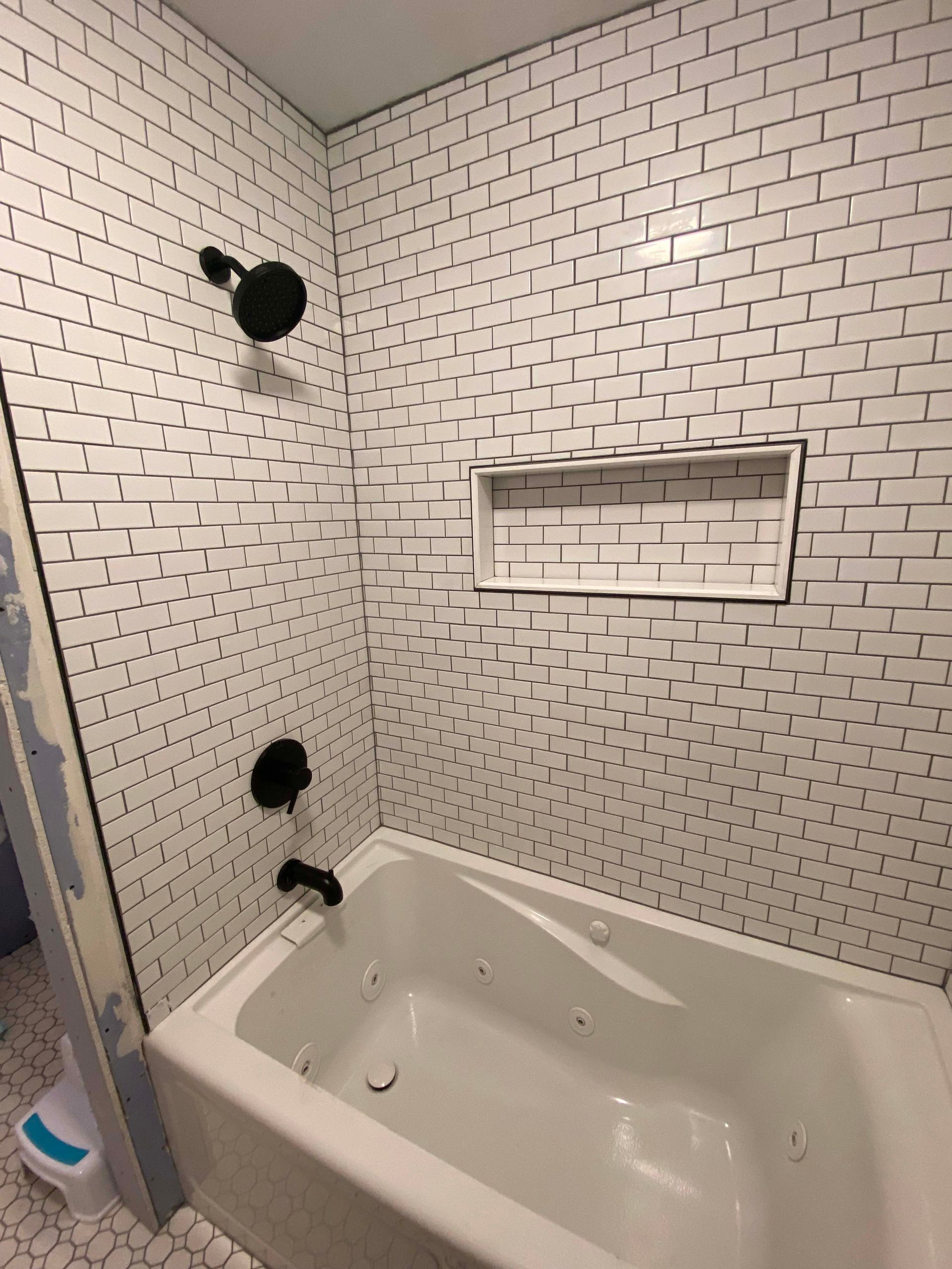 Small tiled bathroom shower with white tub, built-in shelf, and black shower fixtures.