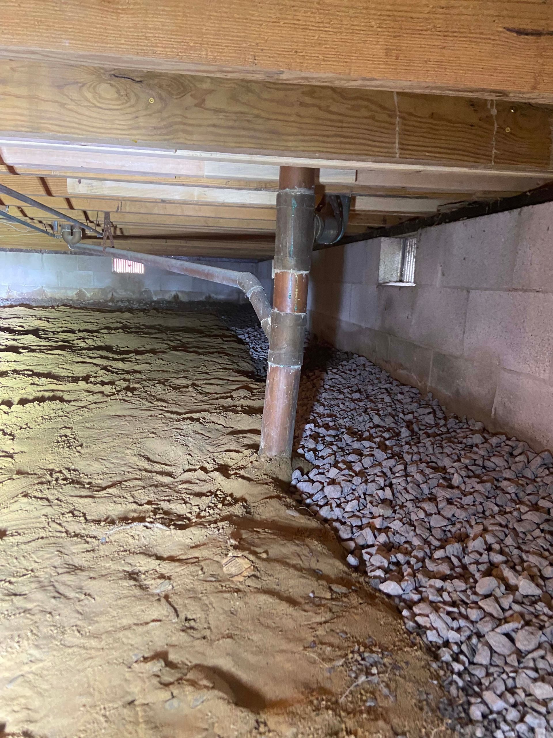 Under-house crawlspace with soil floor, insulation, and exposed plumbing pipe under wooden joists
