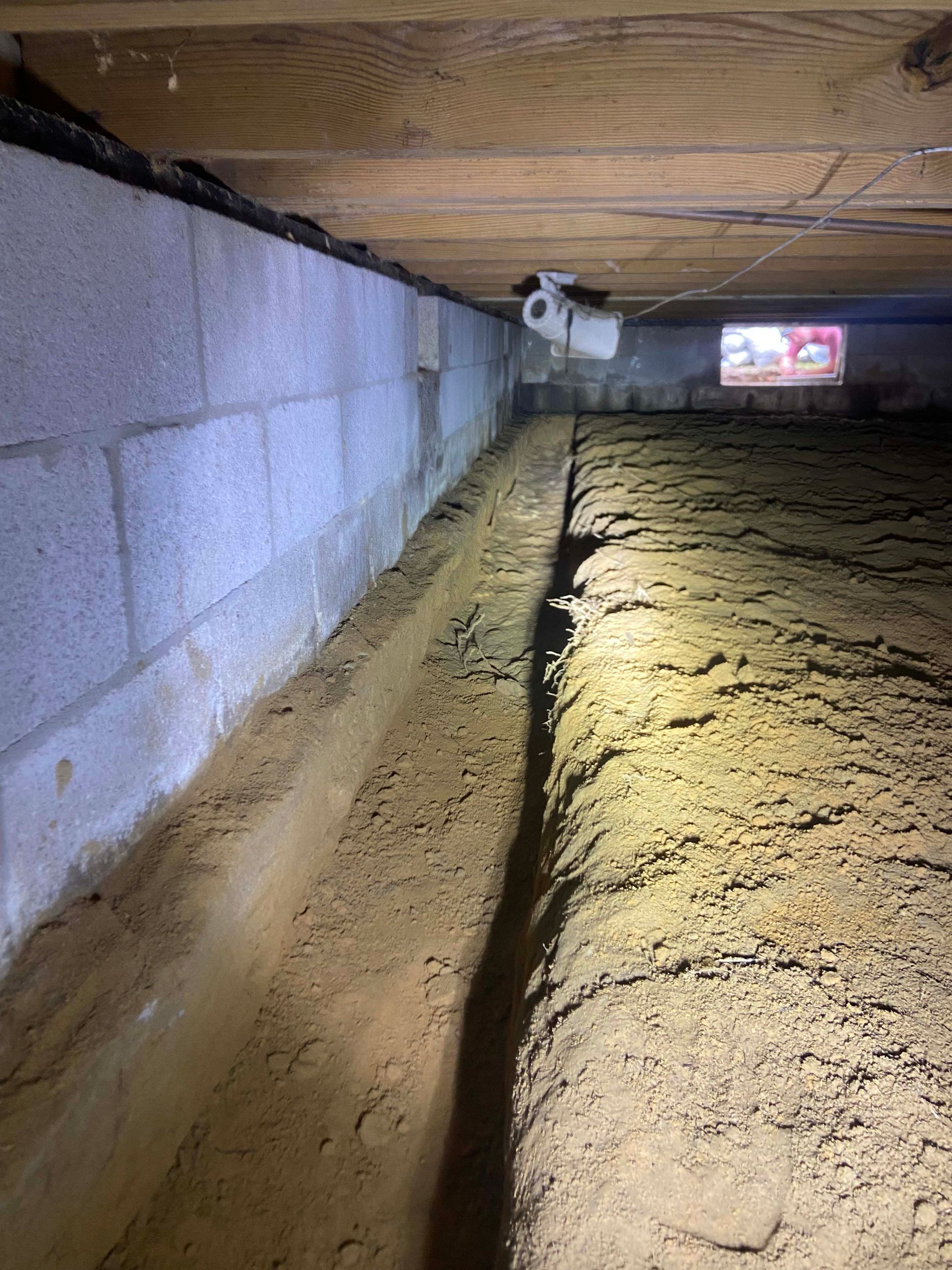 Insulated crawlspace with a narrow dirt trench and bright light shining toward a vent opening
