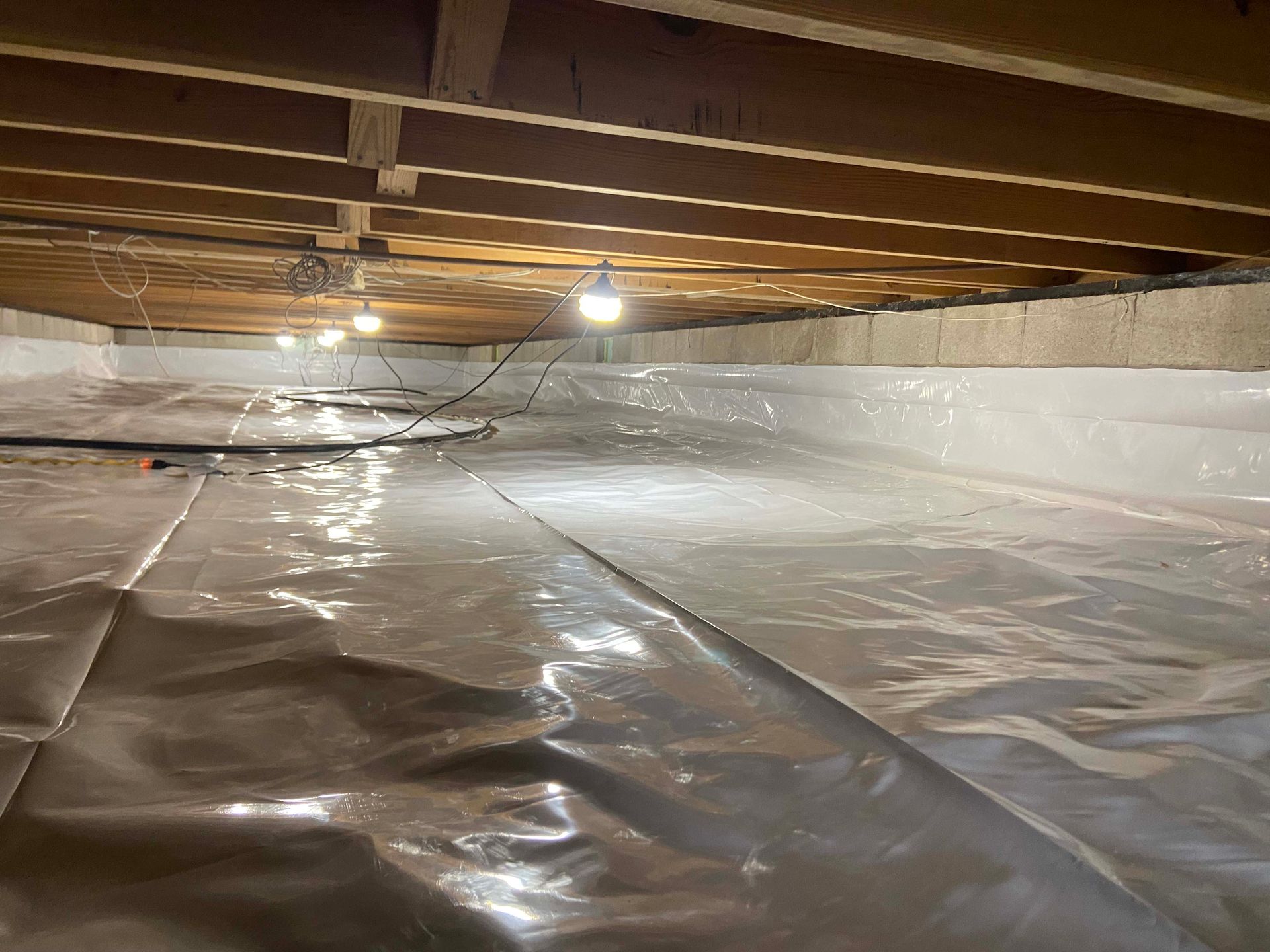 Basement crawlspace with exposed beams and plastic-covered floor, lit by a few bulbs
