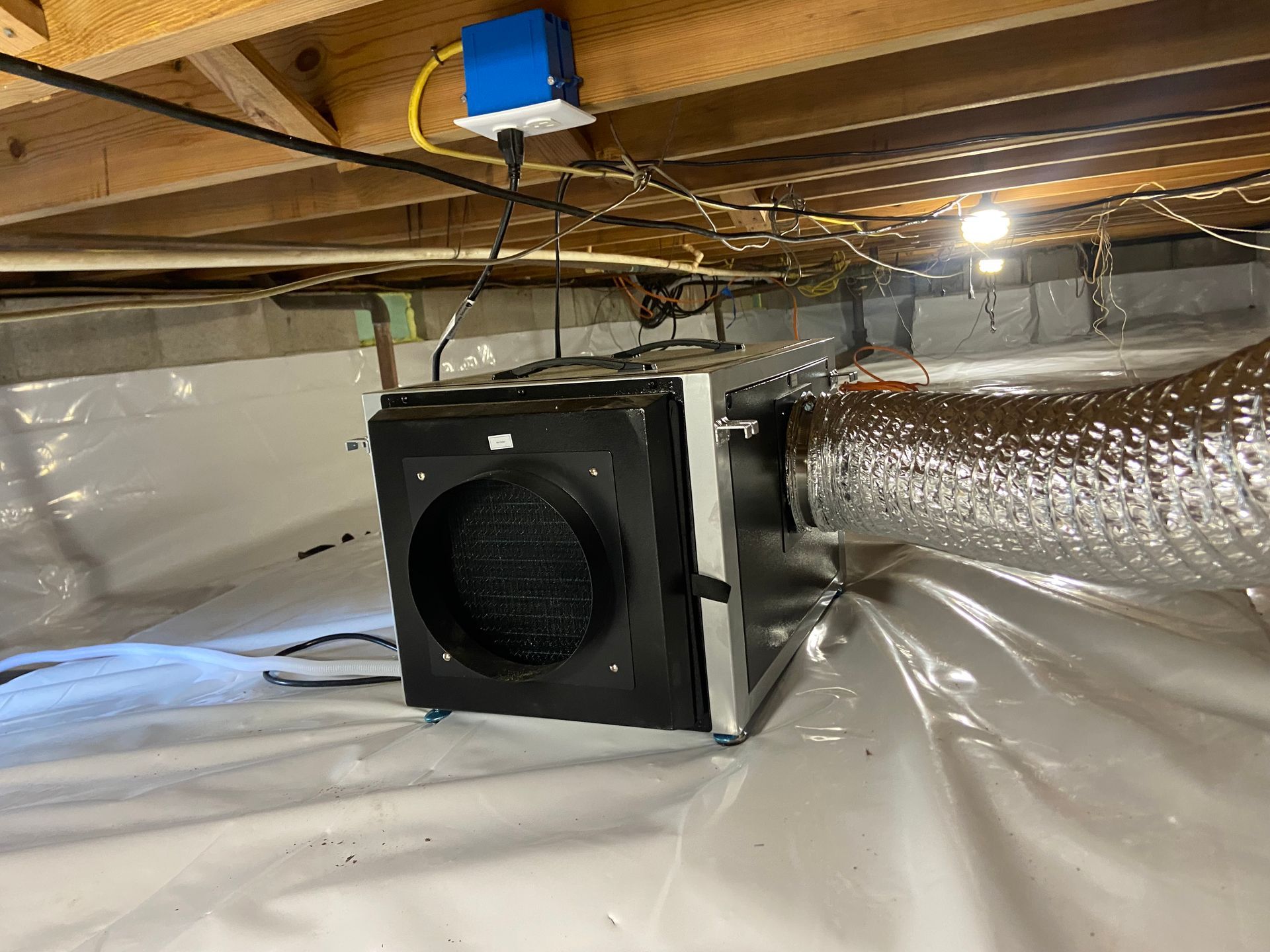 A black ventilation fan under floor joists, connected to silver ducting in a crawlspace.