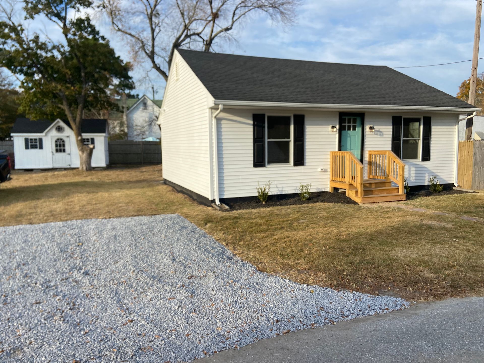 Small beige house with black shutters, turquoise door, and a gravel driveway in front.