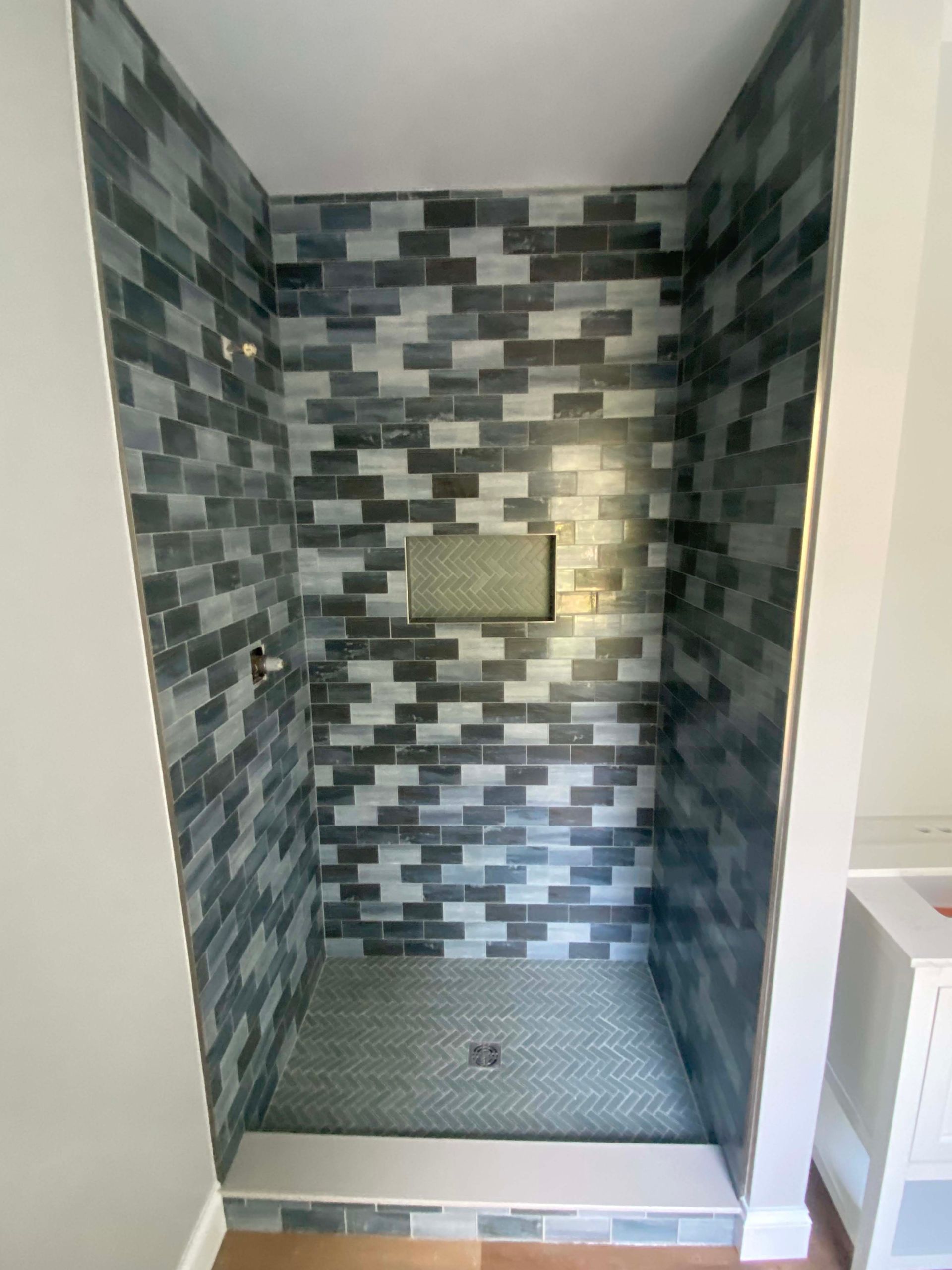 Small tiled shower stall with a drain, built-in shelf, and patterned gray-blue mosaic walls