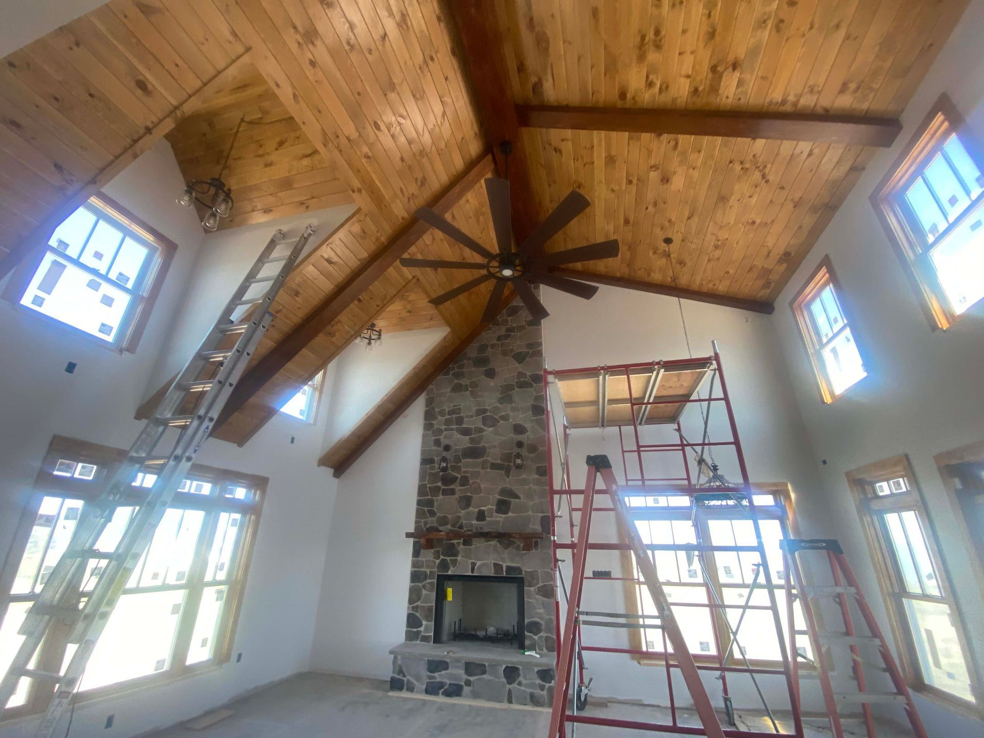 High-ceiling living room with wood plank ceiling, stone fireplace, and ladders/scaffolding during renovation