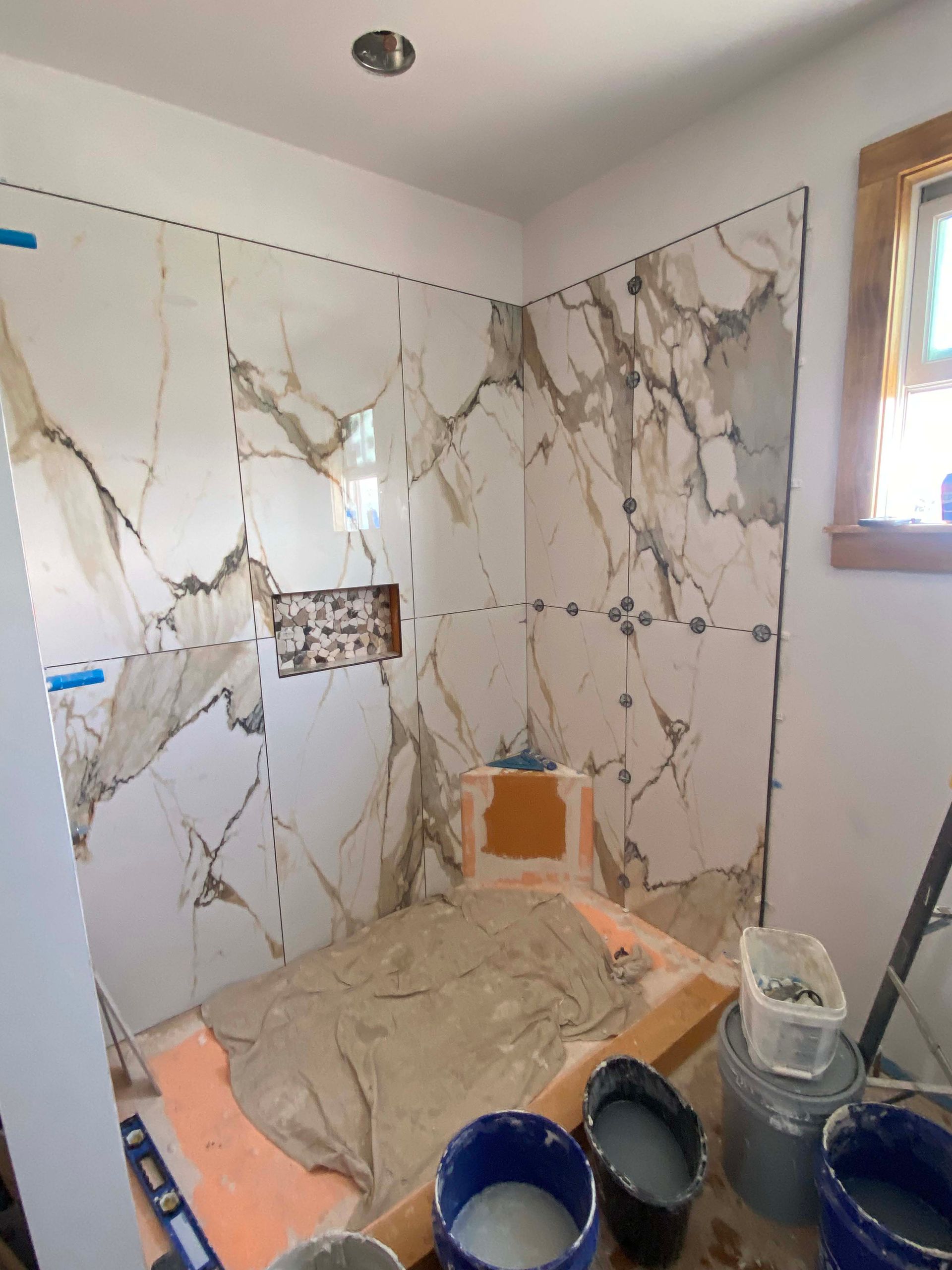 Tiled bathroom shower under renovation, with marble-look wall panels, construction dust, and paint buckets on the floor