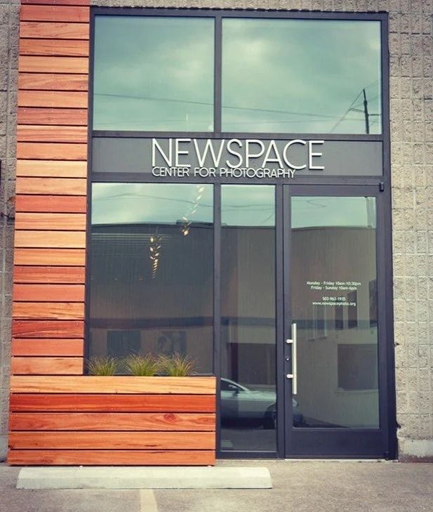 Entrance to Newspace Center for Photography with glass door and windows, wood siding, and signage.