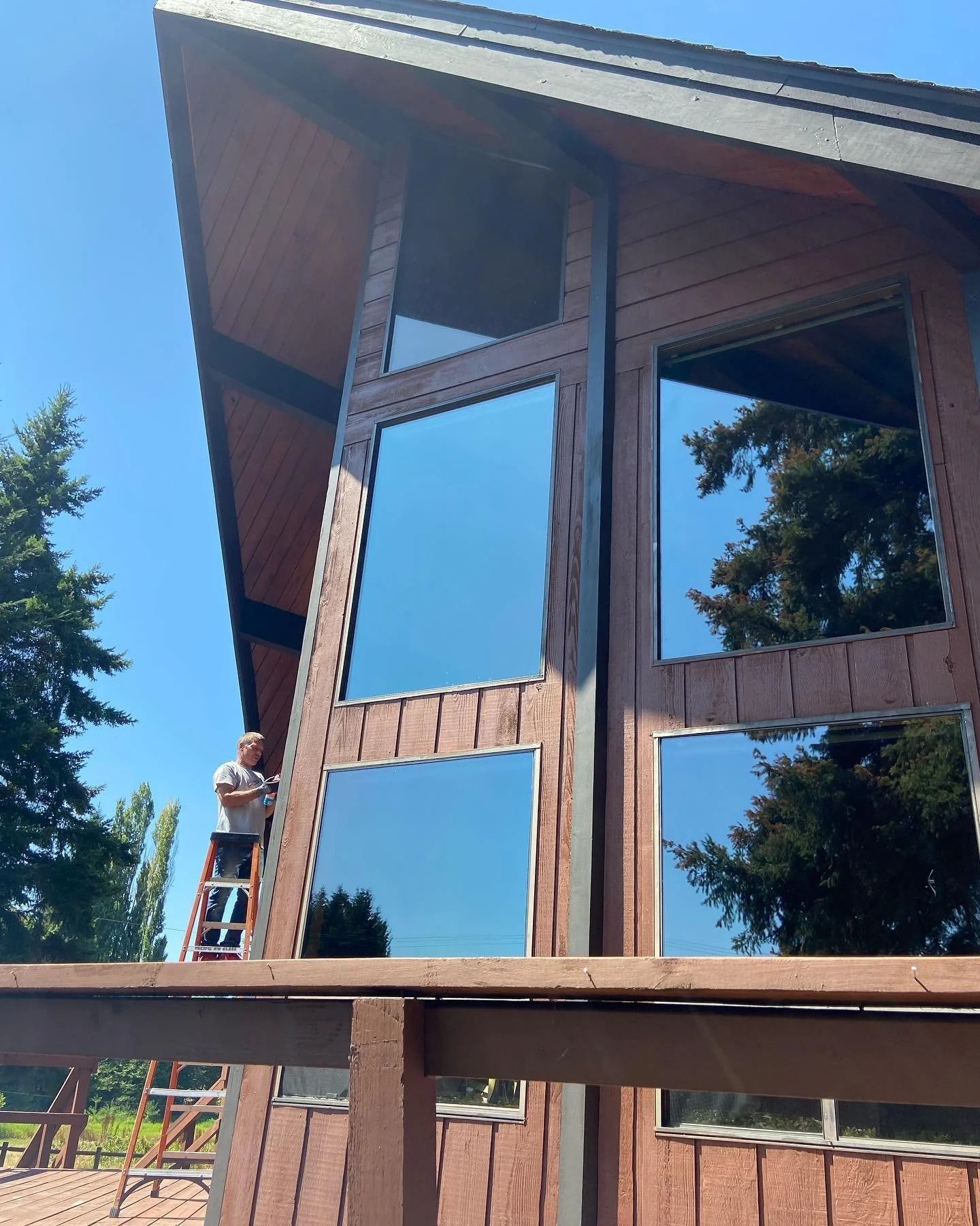Person on ladder cleaning large windows on a brown A-frame house; blue sky reflects in the glass.