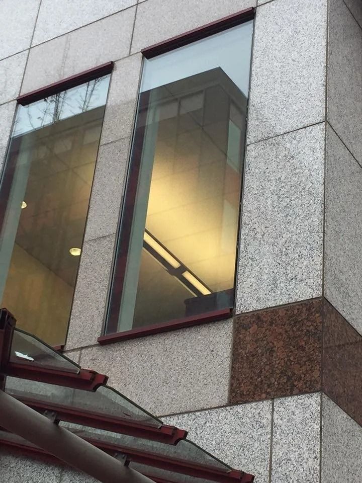 Exterior of a building with granite and glass windows; a reflection shows an interior with fluorescent lights.