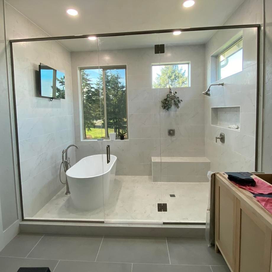 Modern bathroom with glass shower, soaking tub, and windows. Light gray tile, neutral colors.