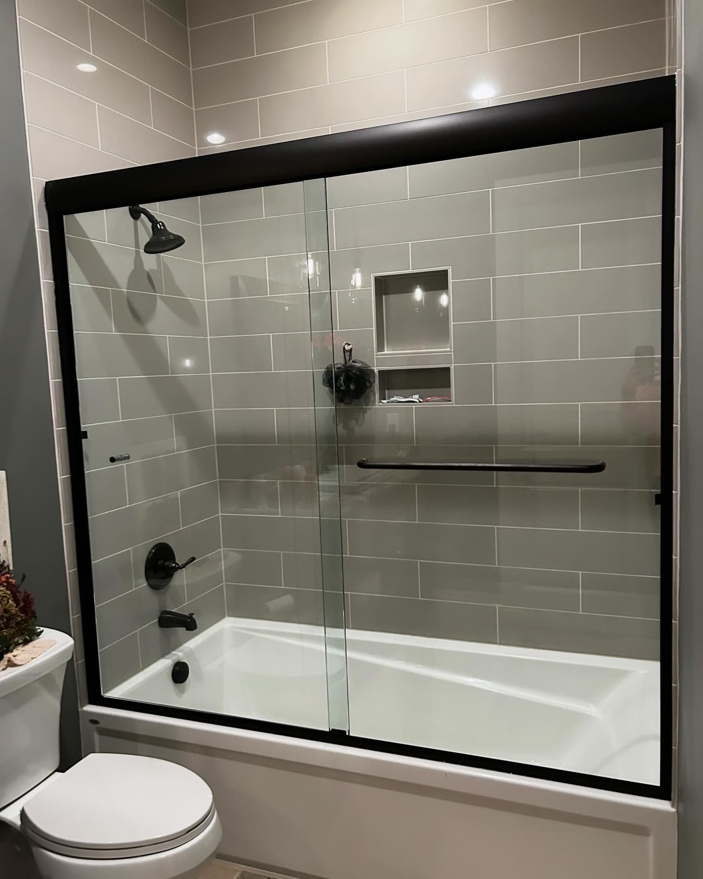 Gray tiled bathroom with a black-framed glass shower door over a white tub.