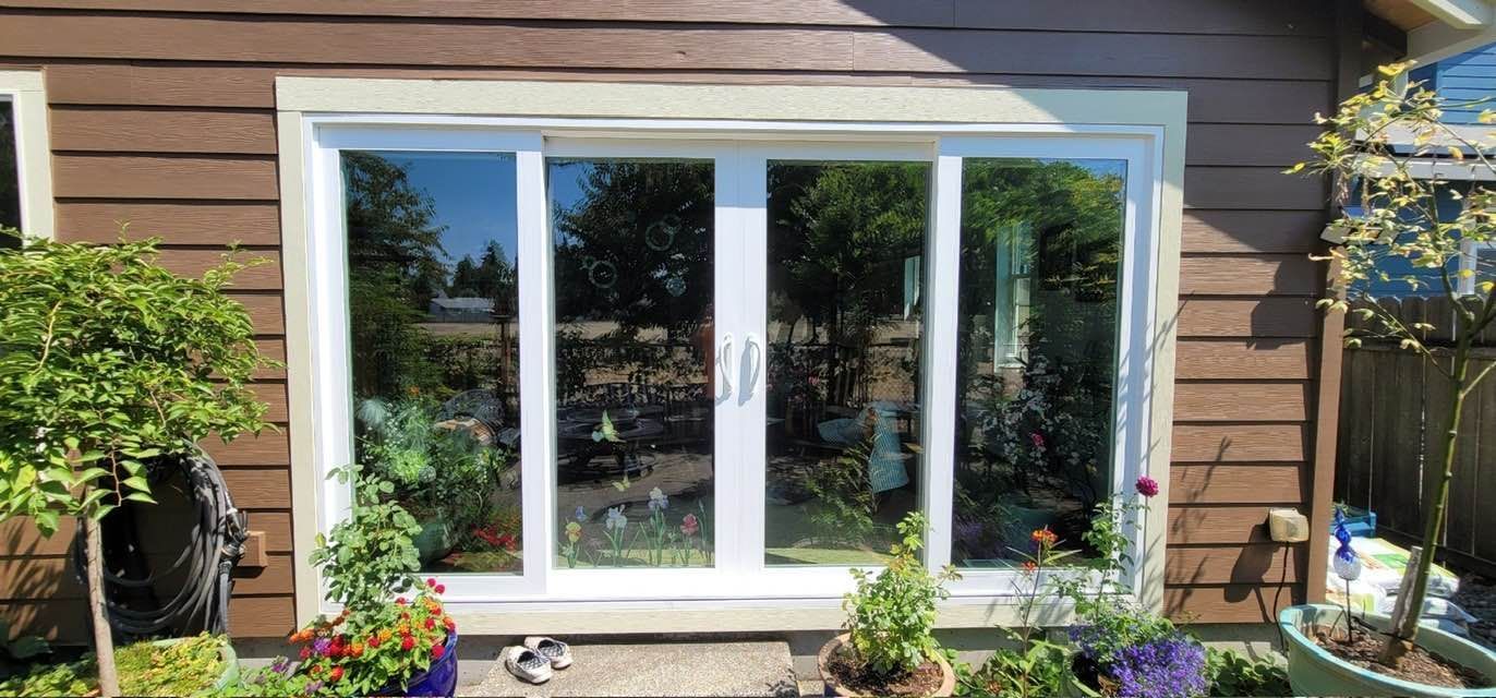 A white framed sliding glass window on a brown wooden house, with plants in the foreground, reflecting the outdoors.