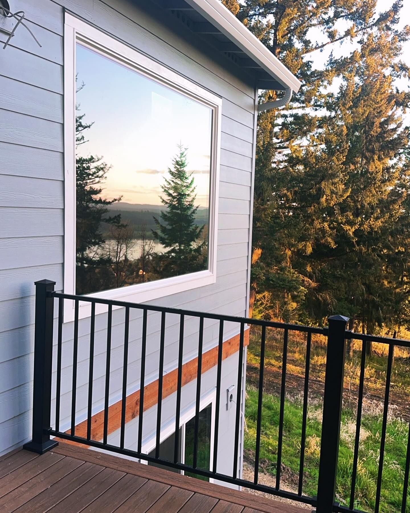 Exterior view of a house with a large window reflecting a tree and landscape. Black railing and siding.