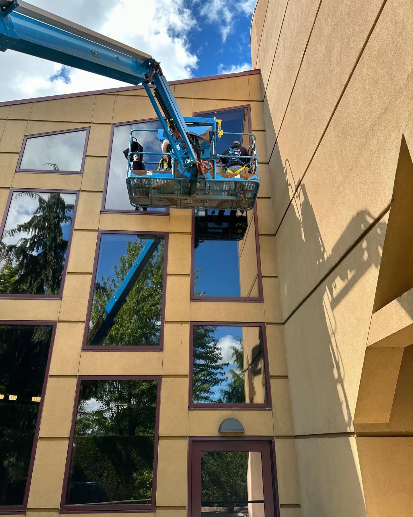 A lift with a worker is cleaning windows on a tan building under a blue sky.