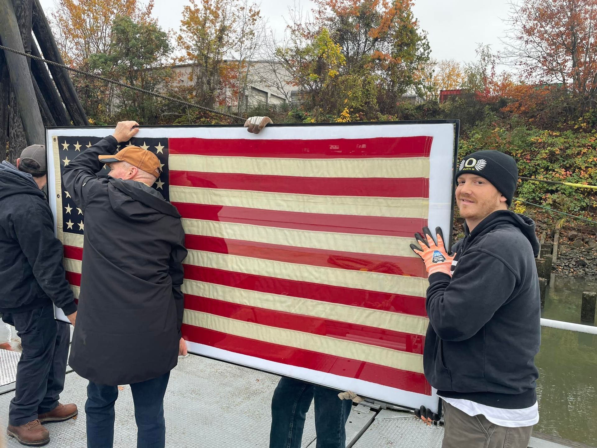 Three people holding a large American flag. Outdoors, cloudy day.