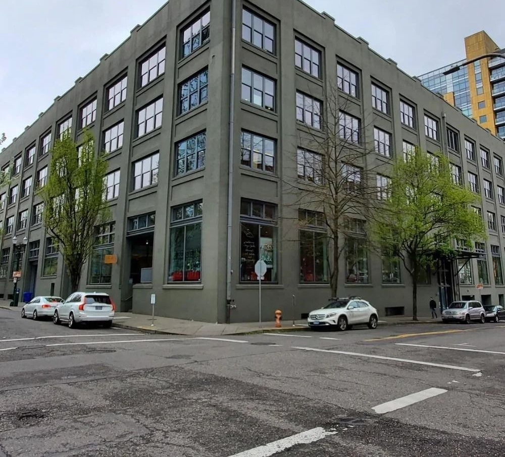 Gray industrial building on corner with cars parked nearby, trees, and cloudy sky.