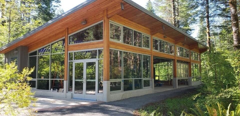 Modern building with large glass windows and wooden beams in a forest setting.
