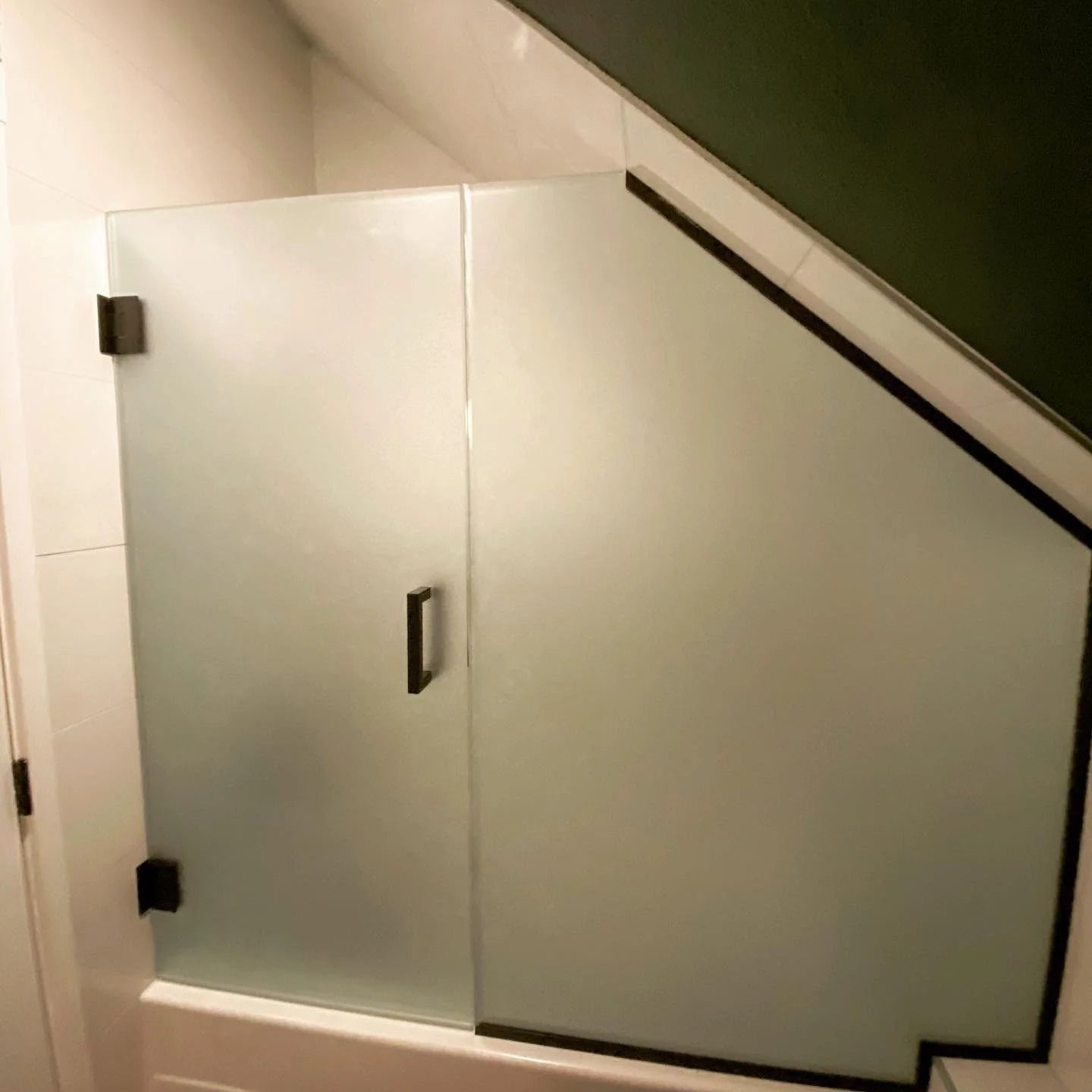 Frosted glass shower door with black hardware, angled section. White tiles and green ceiling.