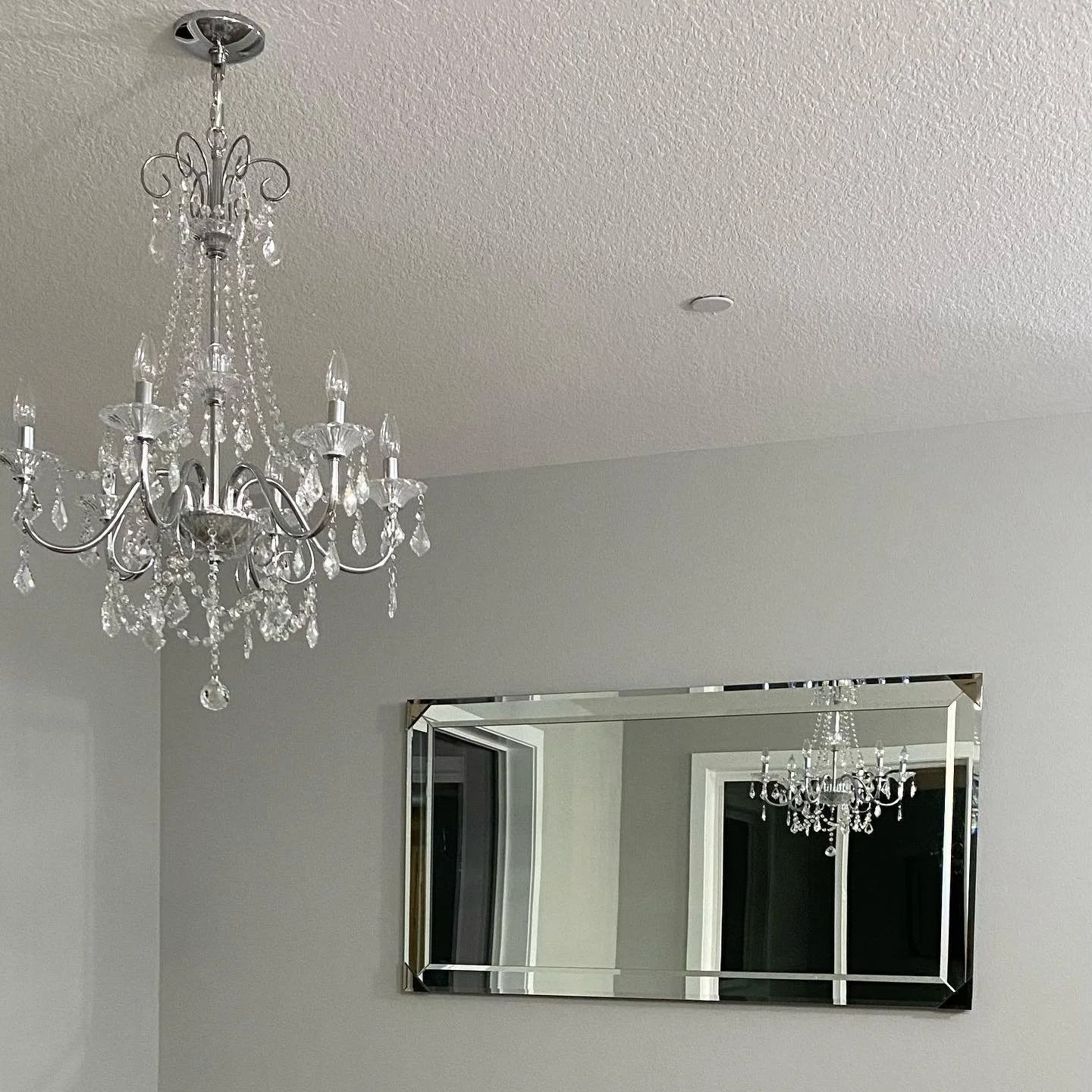 A crystal chandelier hangs near a mirror on a light gray wall. The mirror reflects the chandelier.