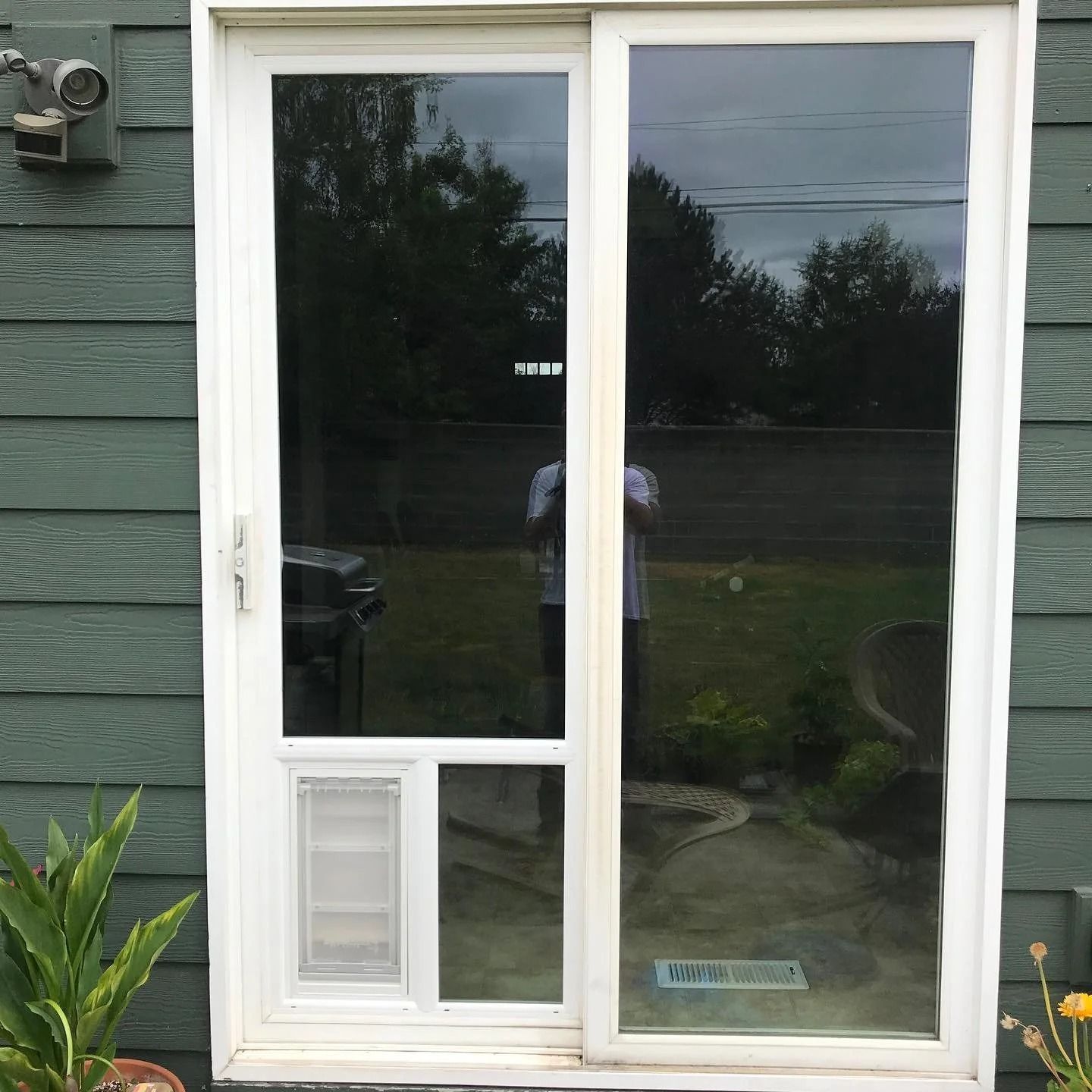 White sliding glass door with pet door, reflecting a yard with trees and person.