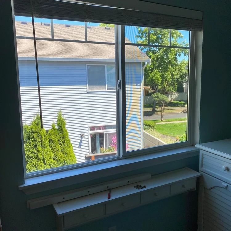 Window view of a blue-sided building, trees, and greenery outside. A built-in white shelf is below the window.