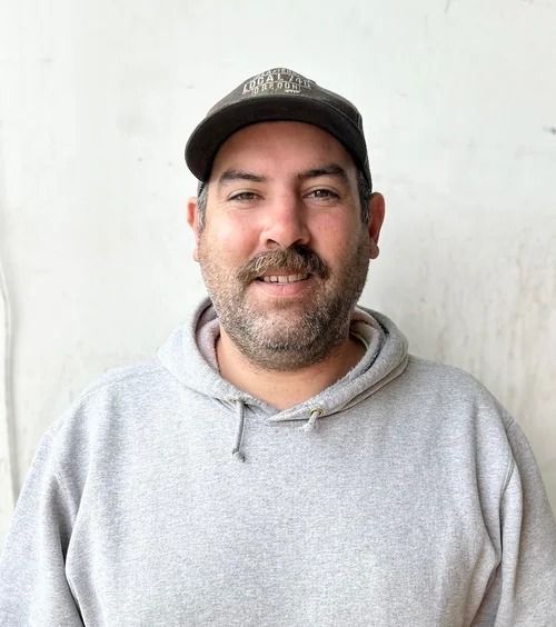 Man in grey hoodie and cap smiles at the camera, standing against a white wall.