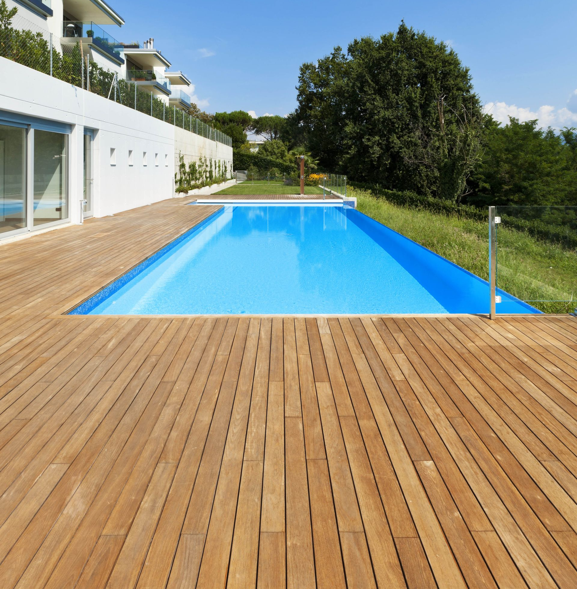 Modern backyard pool with wooden deck, white house, and green lawn under a clear blue sky