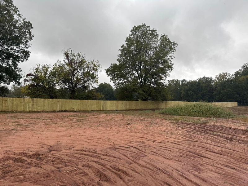 Wooden fence bordering a field with reddish-brown dirt under an overcast sky, trees in the background.