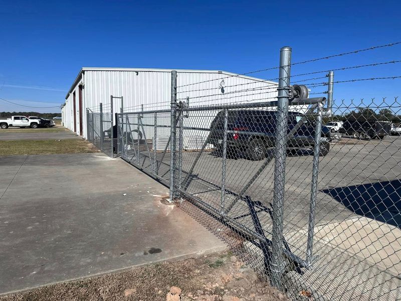 Chain link fence with barbed wire surrounds a gray building; a gate is open with a black vehicle visible.