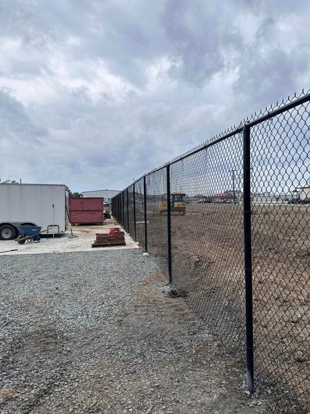 Black chain-link fence bordering a gravel lot, with a trailer and construction equipment visible on the left and in the distance.