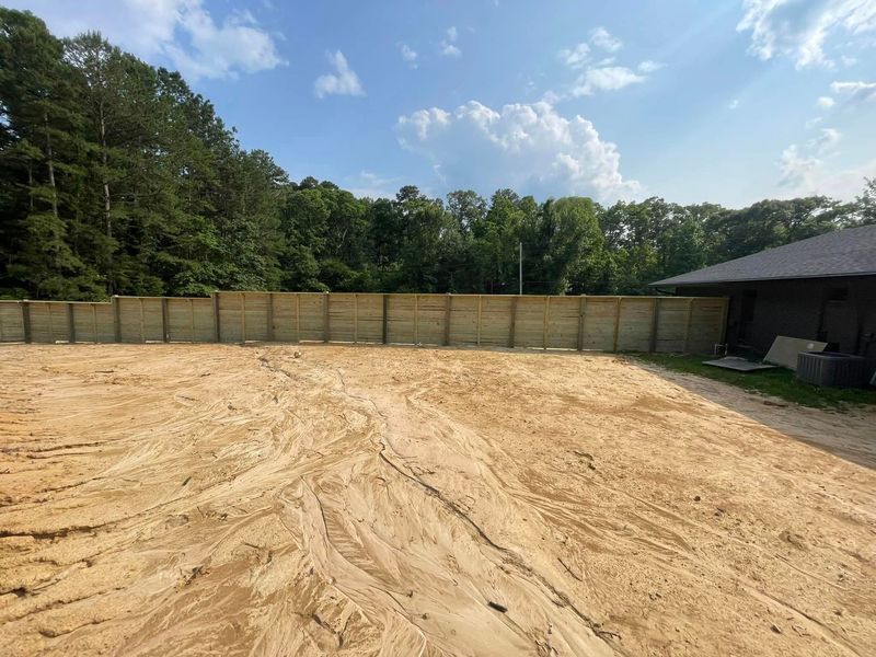A large sandy backyard with a tall wooden fence, trees, and partly cloudy sky.