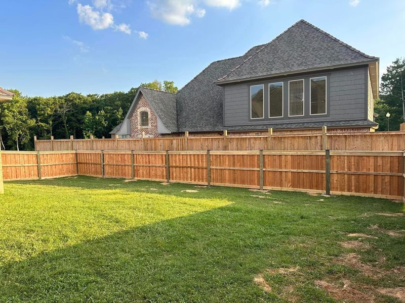 Wooden fence encloses a green lawn, backdrop of a house with gray siding and roof under a blue sky.
