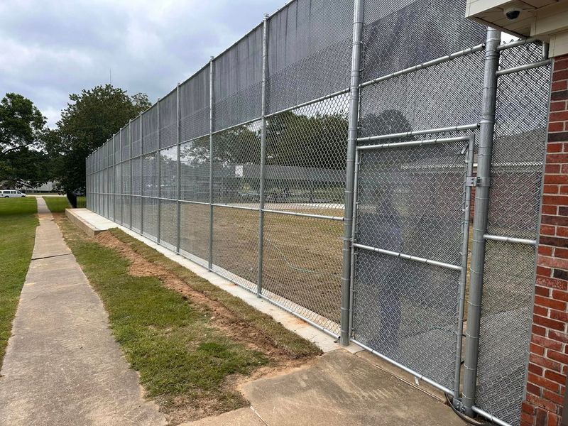 Chain-link fence with a gate along a concrete walkway next to a grassy area, brick building on the right.