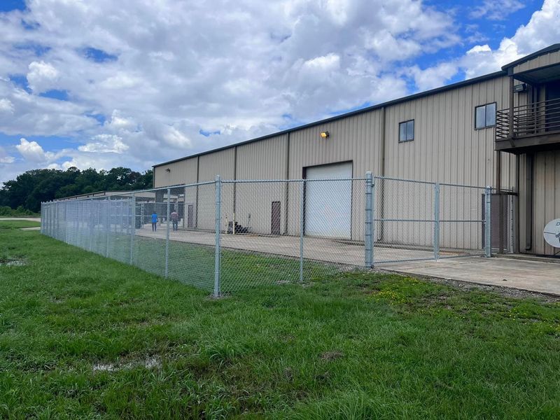 Chain-link fence surrounds a beige industrial building with a white door, set in a grassy area under a cloudy sky.