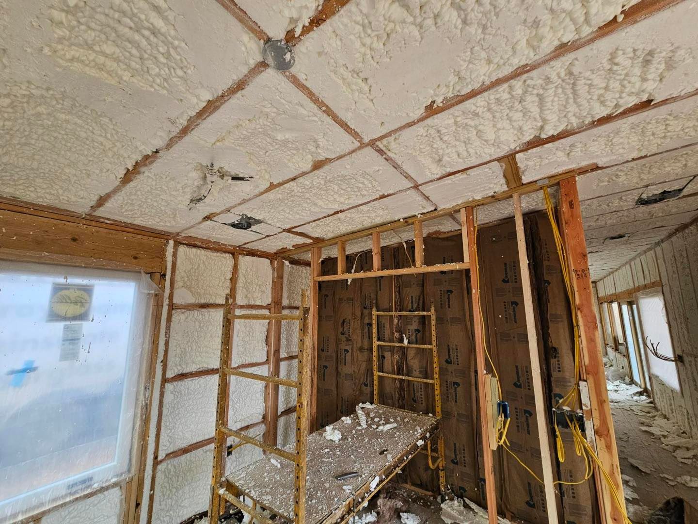 Interior view of a room under construction with spray foam insulation on the walls and ceiling, scaffolding present.