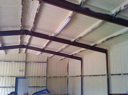 Interior of a metal building insulated with light yellow spray foam, covering walls and ceiling, with brown metal beams.