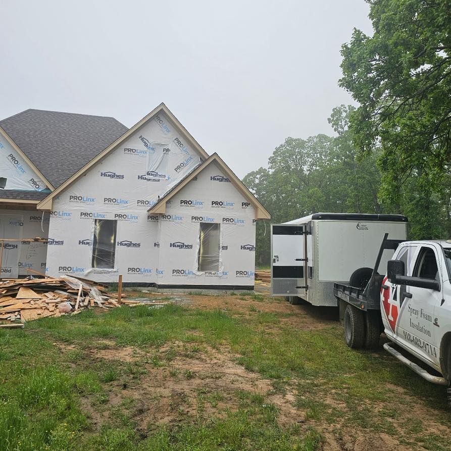 House under construction with white siding, trailer, and truck on a grassy lot. Overcast sky.