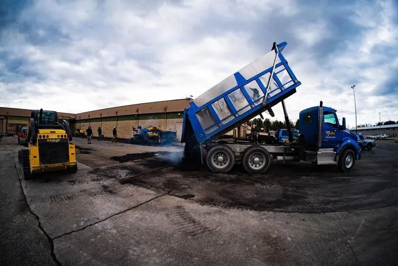 Dump truck unloading asphalt onto cracked pavement; yellow skid steer loader nearby.