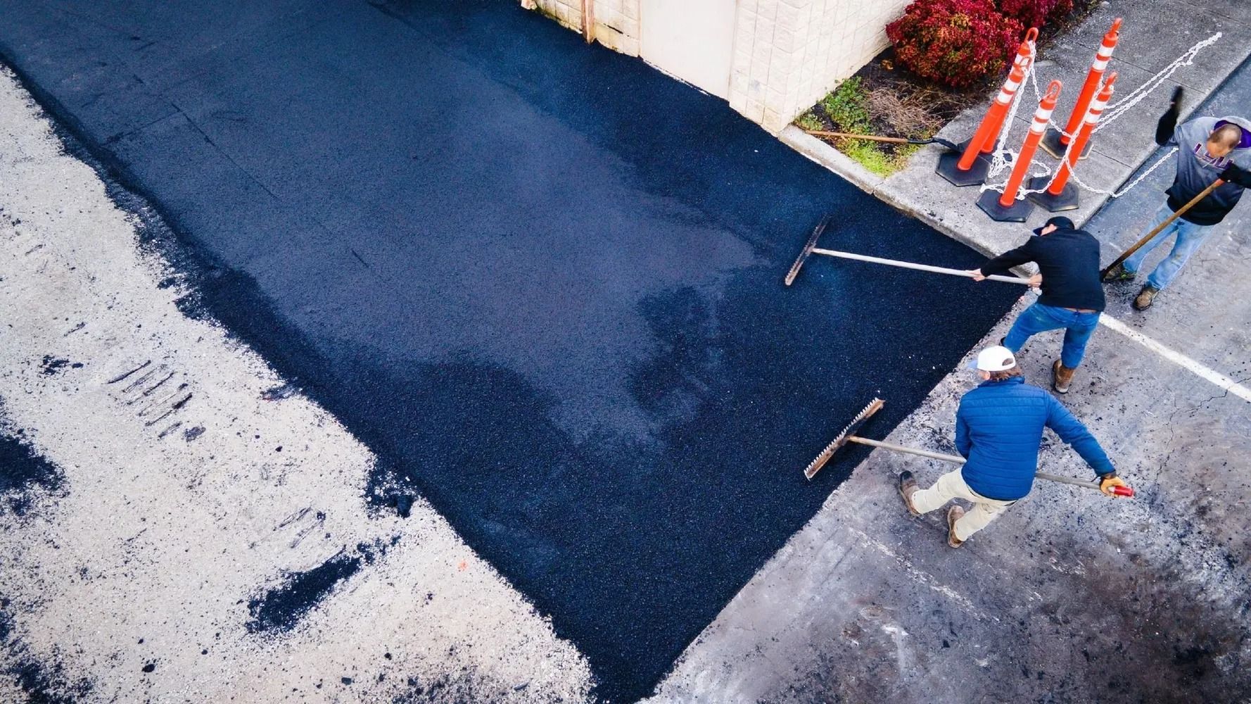 Workers paving a road, using rakes to spread the dark asphalt. Orange cones are present in the setting.