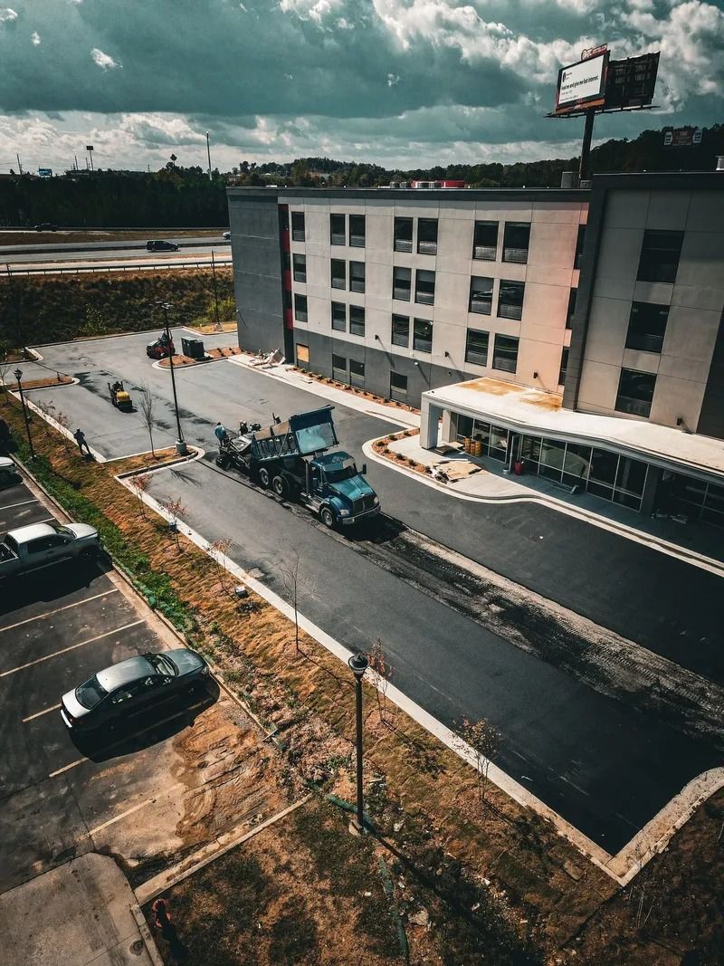 Aerial view: Construction truck paving a hotel driveway; cars parked in the lot; overcast sky.