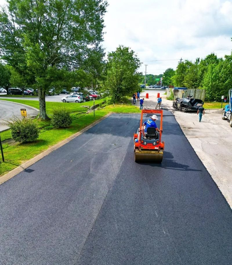 Worker operating a road roller on freshly paved asphalt. Green trees and parked cars in the background.