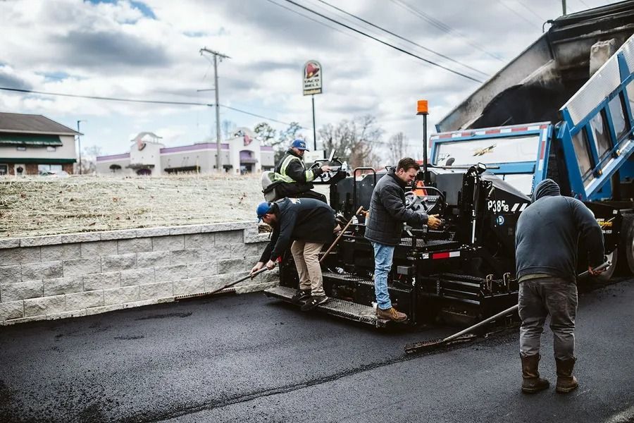 Road workers paving a street with an asphalt machine; cloudy sky in background.