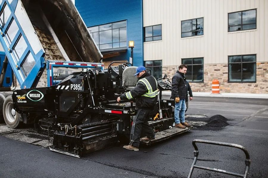 Workers paving a parking lot with asphalt, next to a dump truck and modern building.