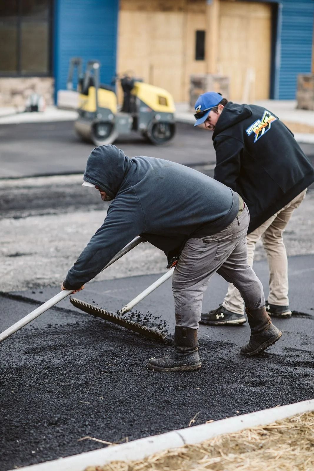Two men paving asphalt road, using rakes. One wears a hood. Yellow roller in background.