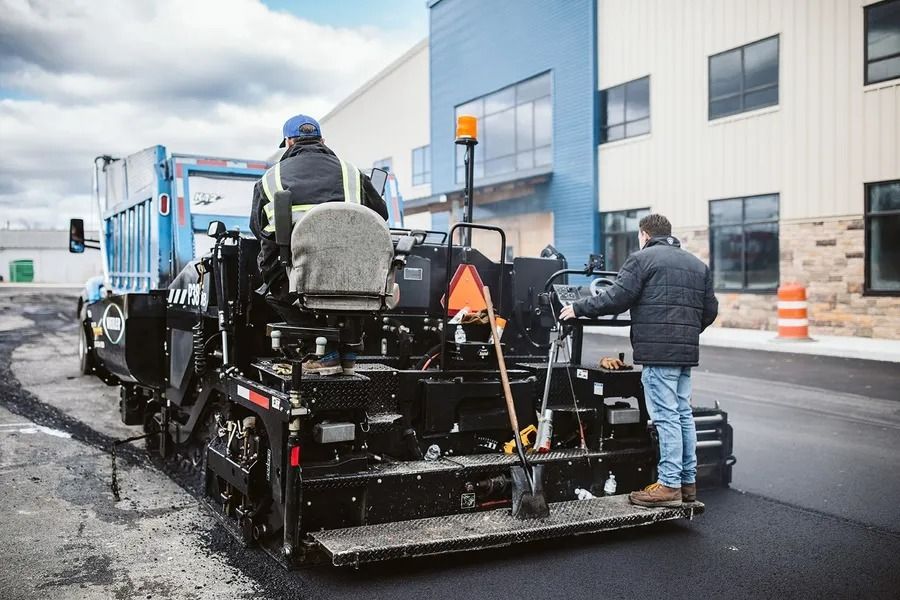 Asphalt paving machine laying black asphalt on a road, with two workers operating it near a building.