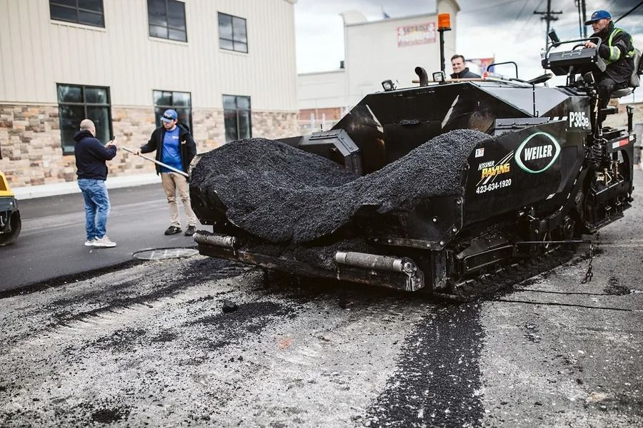 Asphalt paving machine laying new road surface; workers observing.
