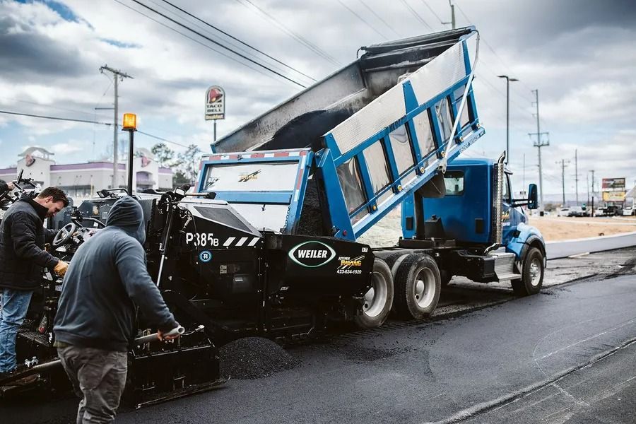 Workers paving a road with a dump truck unloading asphalt on a cloudy day.