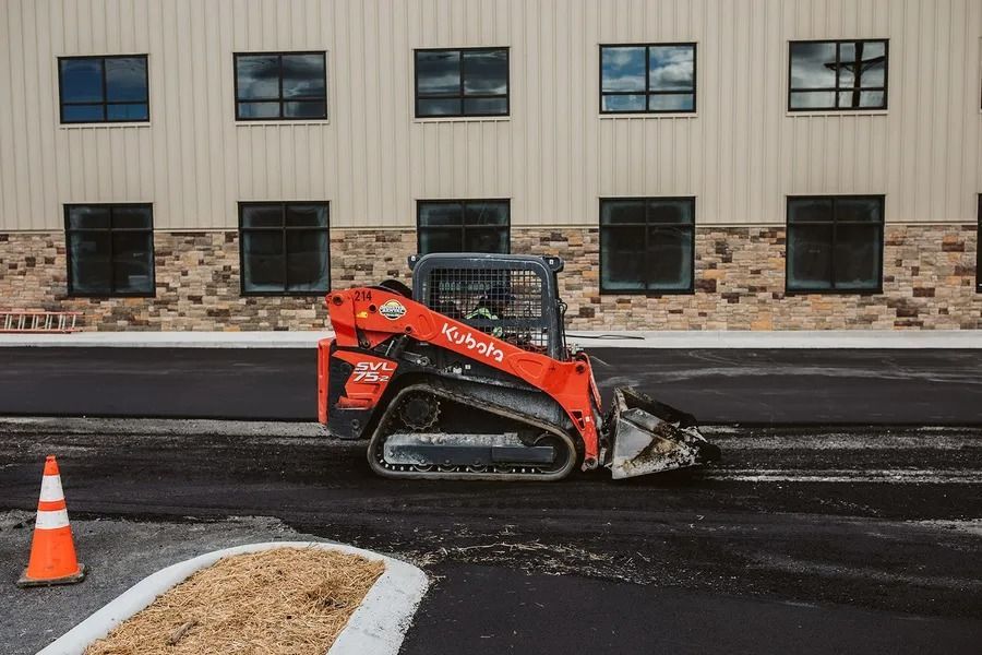 Orange Kubota track loader smoothing asphalt near a building with windows.